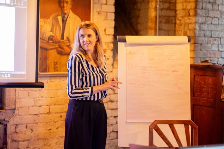 A photo of a woman speaking in front of a flipchart. She has a marker pen in one hand and there is writing on the chart.