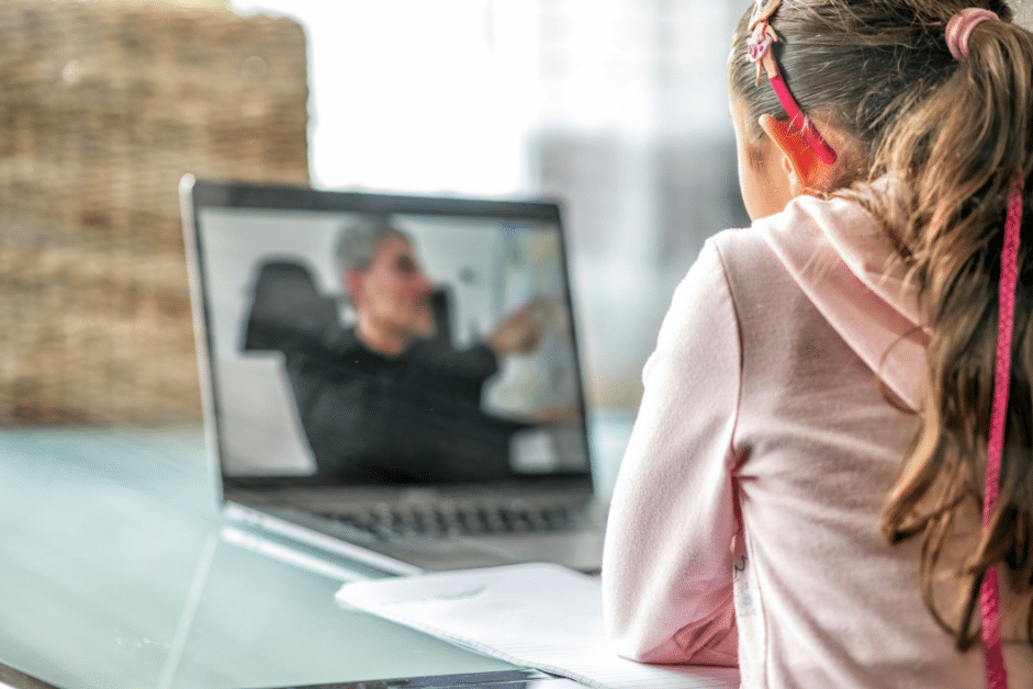 A blurred image of a laptop with a man on the screen demonstrating something. A young girl is sitting at the desk watching the man on the screen.