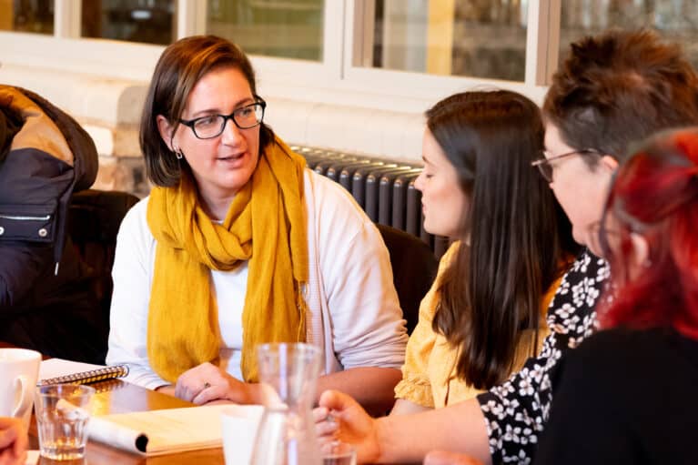 A photo of four women talking together. A notepad with writing on is on the table in front of them.