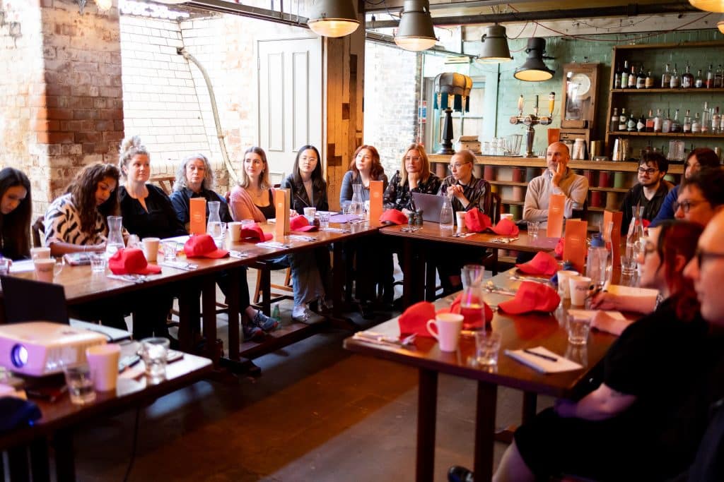 A photo of a room with tables in a U shape. There are red caps on the tables and people are listening to a speaker who is off camera.