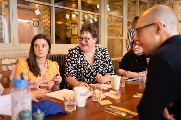 A photo of a group of people sitting round a table covered with notepads, cups and glasses, talking together.