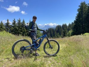 Chris is standing with his bike. In the background, there are trees and a mountain with snow on the top.